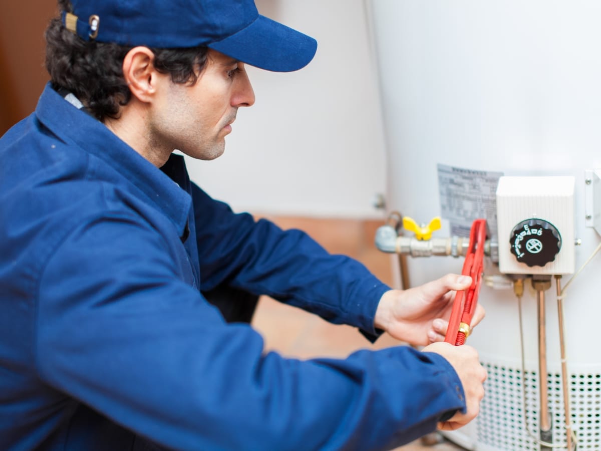 A technician in blue uniform uses a pipe wrench to adjust a valve on a water heater, ensuring everything runs smoothly as part of an annual furnace inspection.