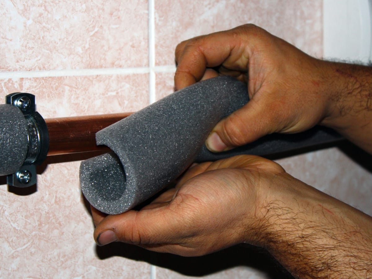 Hands installing foam pipe insulation onto a copper pipe against a tiled wall to help prevent frozen pipes.