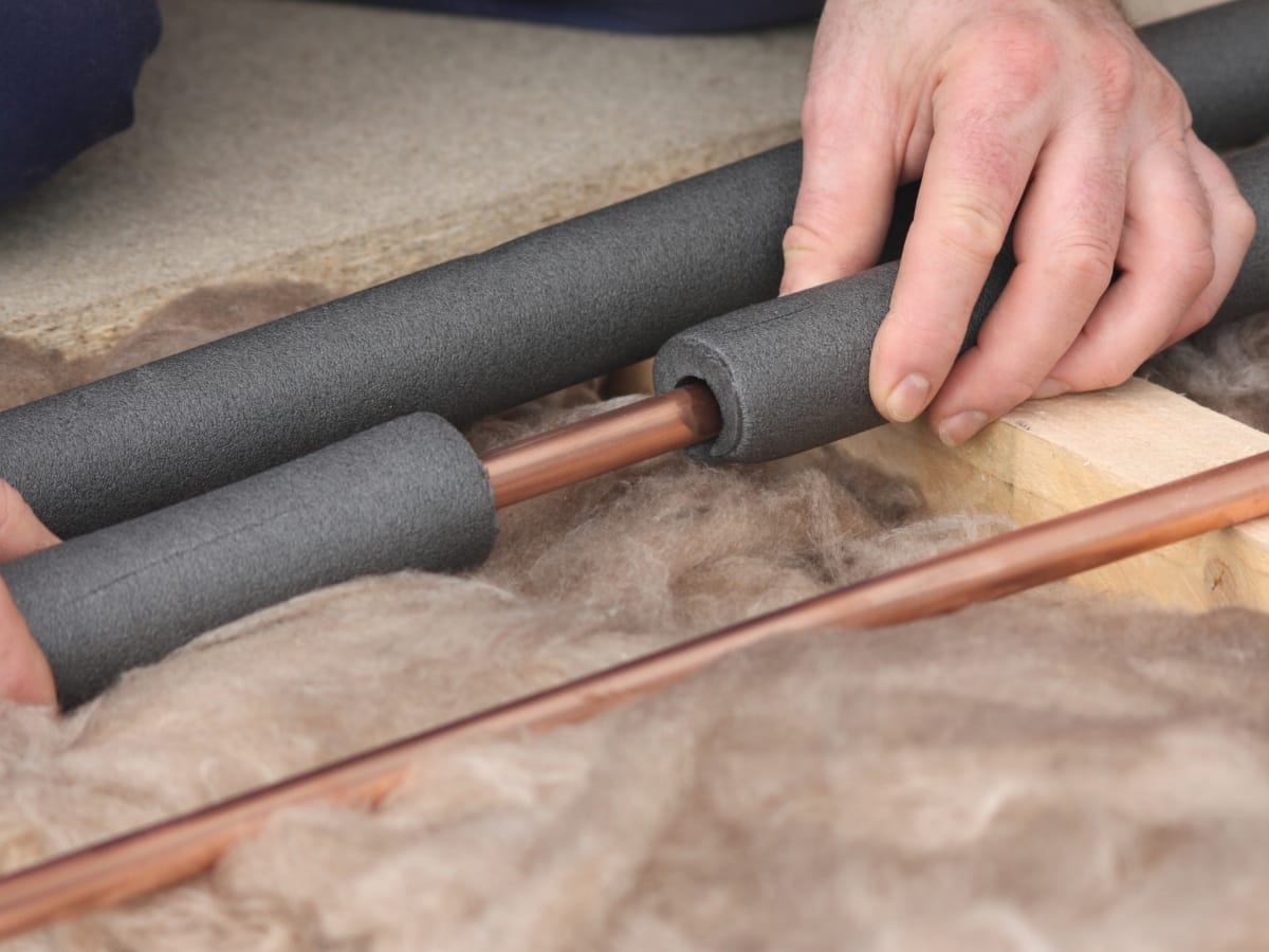 Hands installing foam pipe insulation around a copper pipe with wooden and insulation material in the background to help prevent frozen pipes.