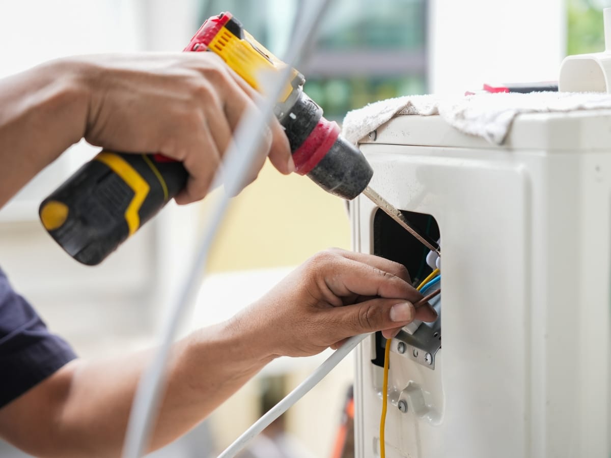A person uses a cordless drill to repair electrical wiring inside an appliance, showcasing Winter Efficiency Tips with various cables and tools visible to ensure safe and efficient operation during colder months.
