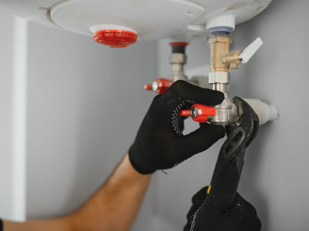 A person wearing black gloves uses a wrench to adjust a valve on a water heater, demonstrating routine boiler services.