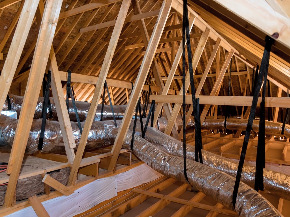 Exposed attic with wooden rafters and beams, featuring metal ductwork and HVAC ducts suspended by black straps along the ceiling structure.