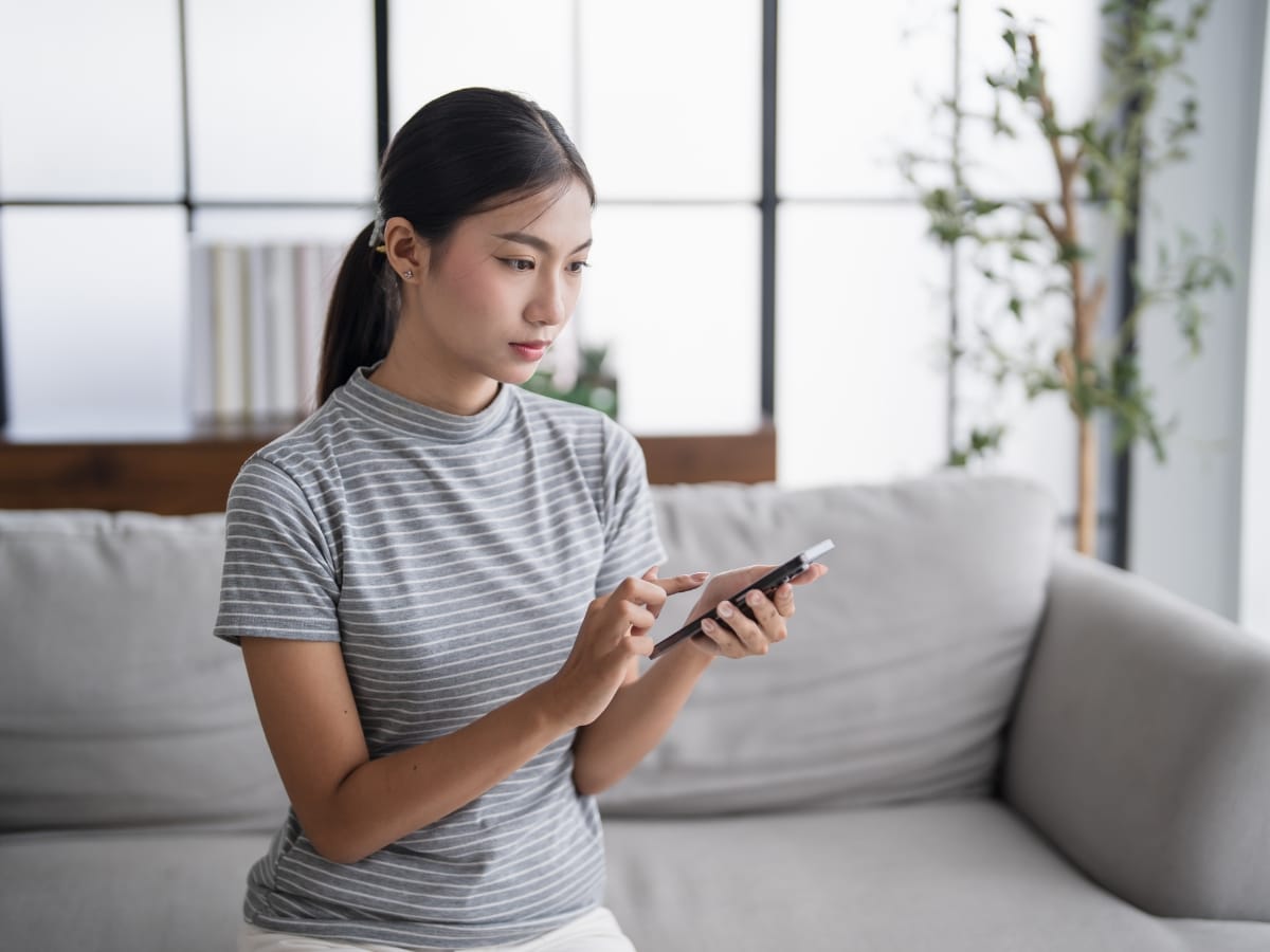 A woman sitting on a gray sofa, using her smartphone with both hands, perhaps exploring smart thermostats benefits. She wears a gray striped shirt and is indoors with a plant in the background.