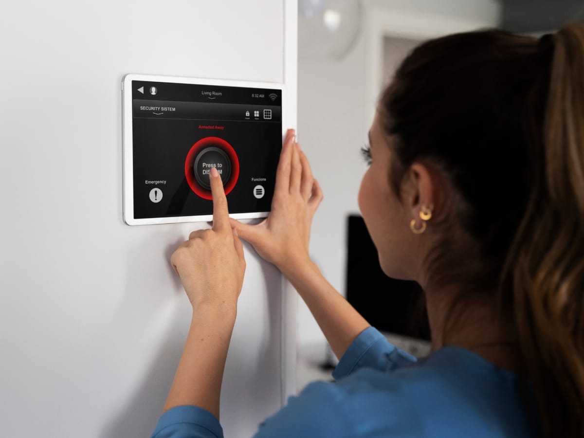 A woman presses a "Press to Disarm" button on a wall-mounted home security touchscreen panel, highlighting home automation features often found alongside smart thermostats benefits.