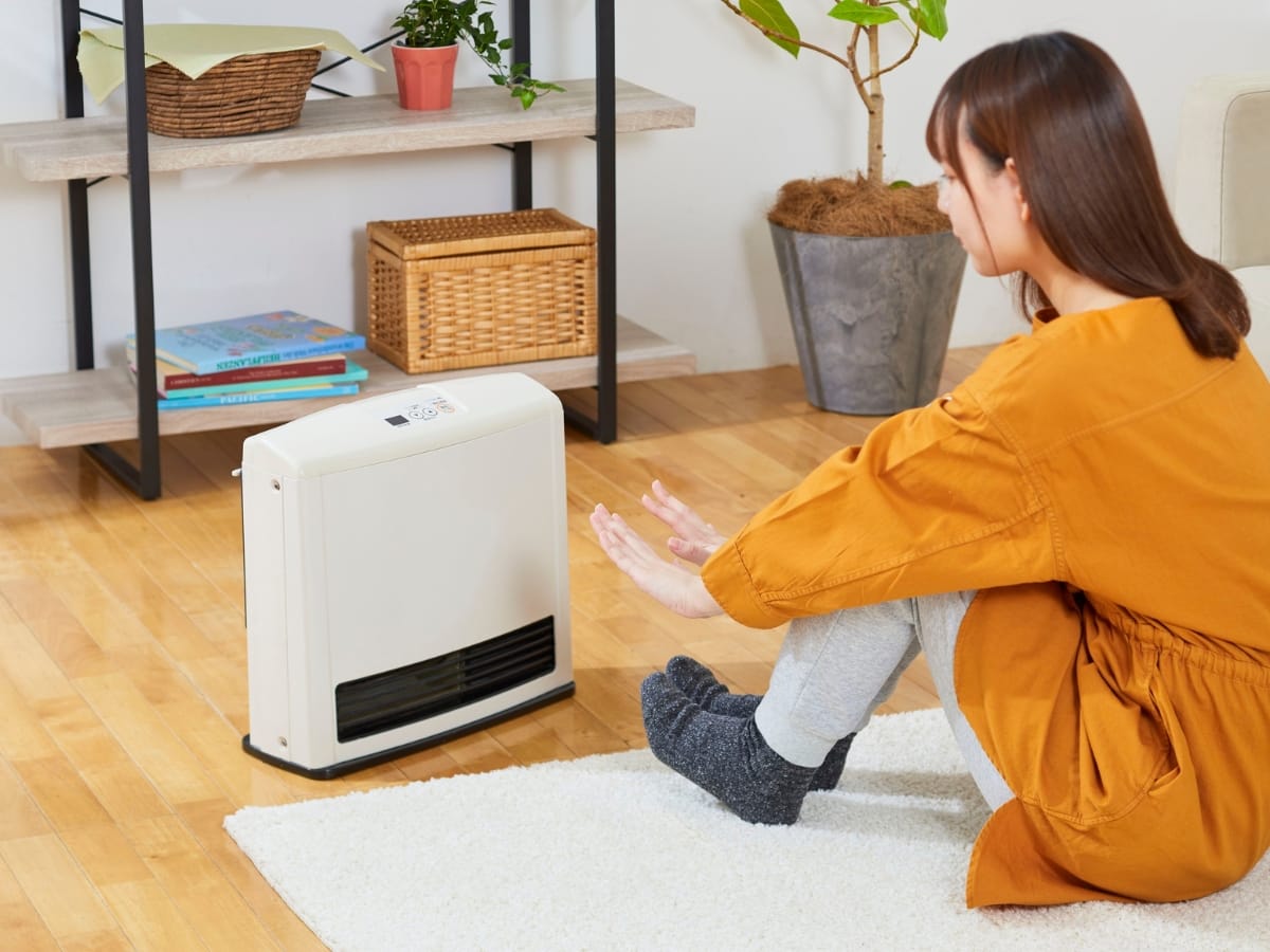 A woman in an orange dress sits on the floor warming her hands in front of a white space heater in a living room, practicing winter heating safety.