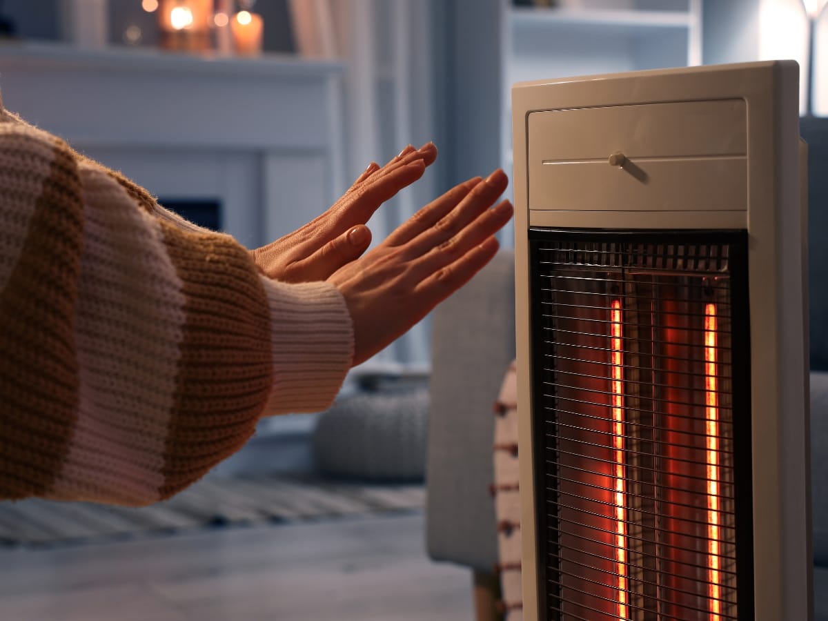 A person in a striped sweater warms their hands in front of an electric space heater, practicing winter heating safety in a cozy indoor setting.