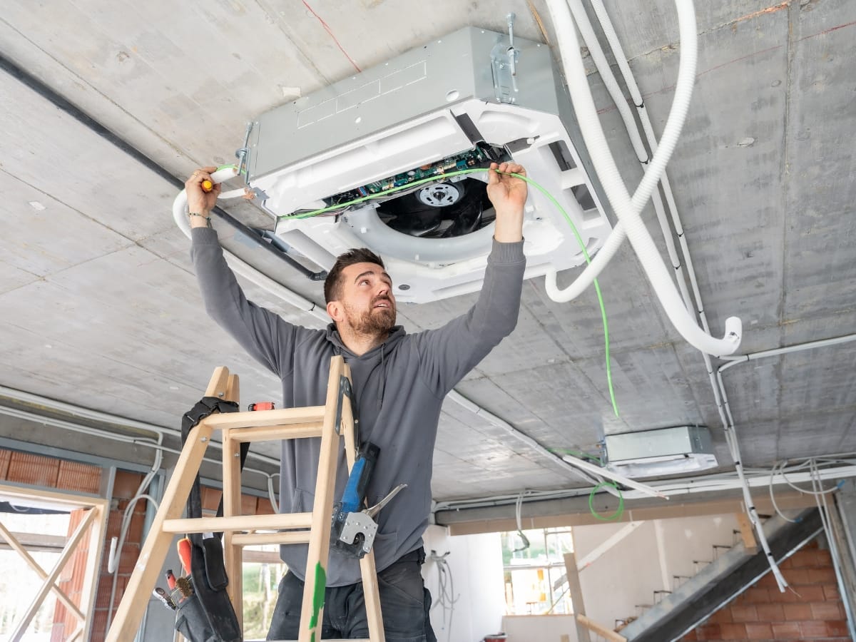 A man standing on a ladder installs or repairs a ceiling-mounted air conditioning unit in an unfinished building, ensuring proper ventilation to help prevent carbon monoxide buildup.