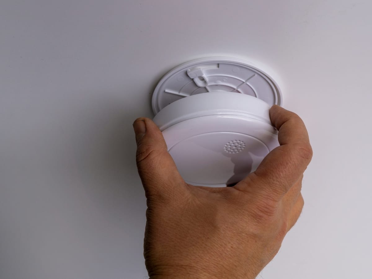 A person’s hand is installing or removing a round smoke and carbon monoxide detector from a white ceiling.
