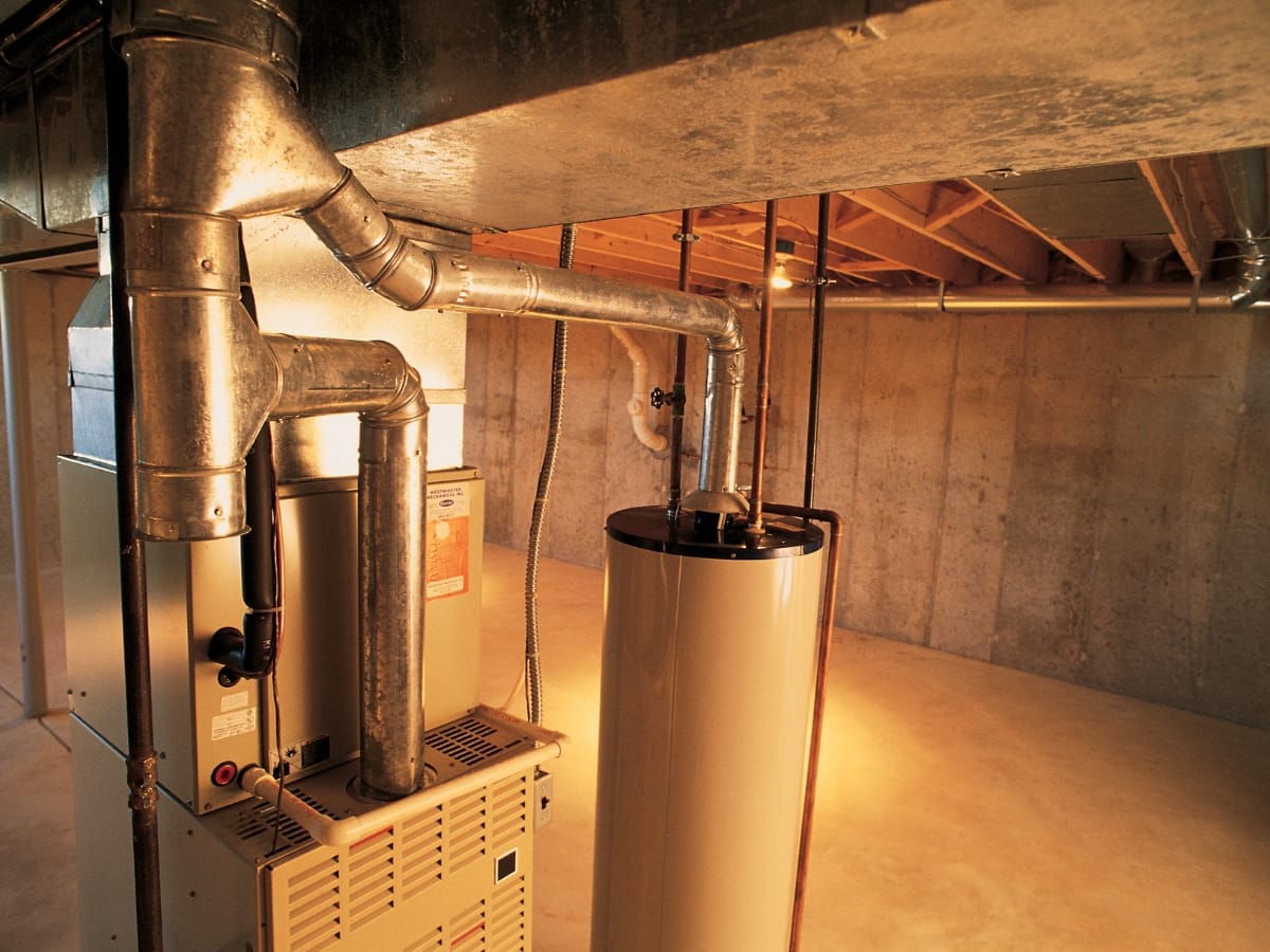 Heating furnace and water heater installed in an unfinished basement with concrete walls and exposed ceiling beams.