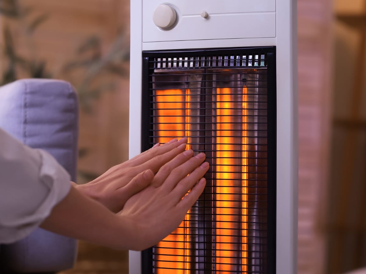 A person warms their hands in front of an electric space heater with glowing heating elements, enjoying the comforting heating effect on a chilly day.