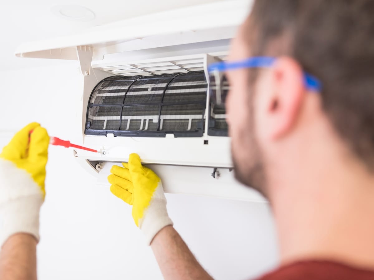 A person wearing yellow gloves uses a screwdriver to repair or perform AC filter cleaning on a wall-mounted air conditioning unit.