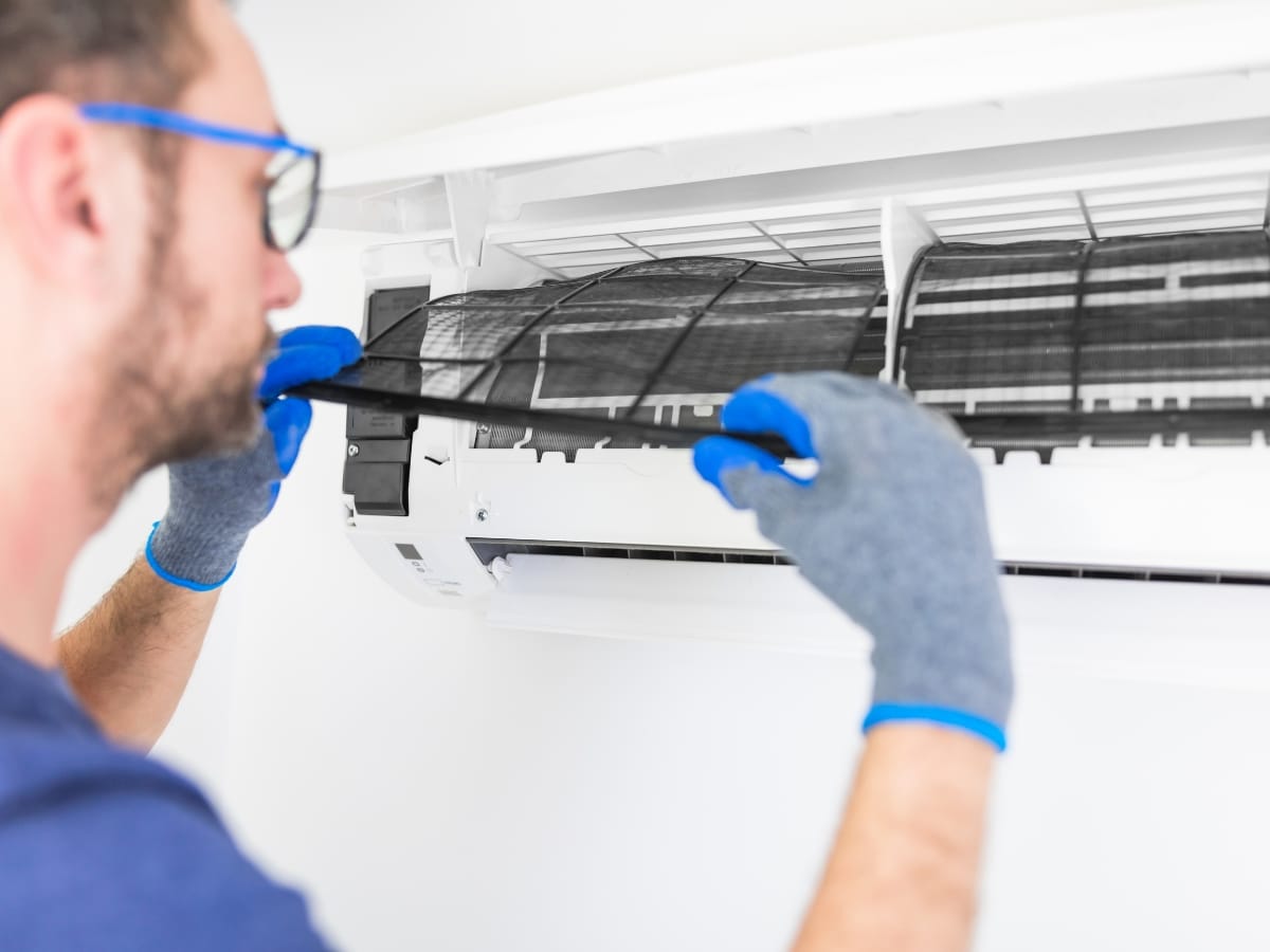 A person wearing gloves is performing AC filter cleaning by removing or cleaning the filter of a wall-mounted air conditioning unit.