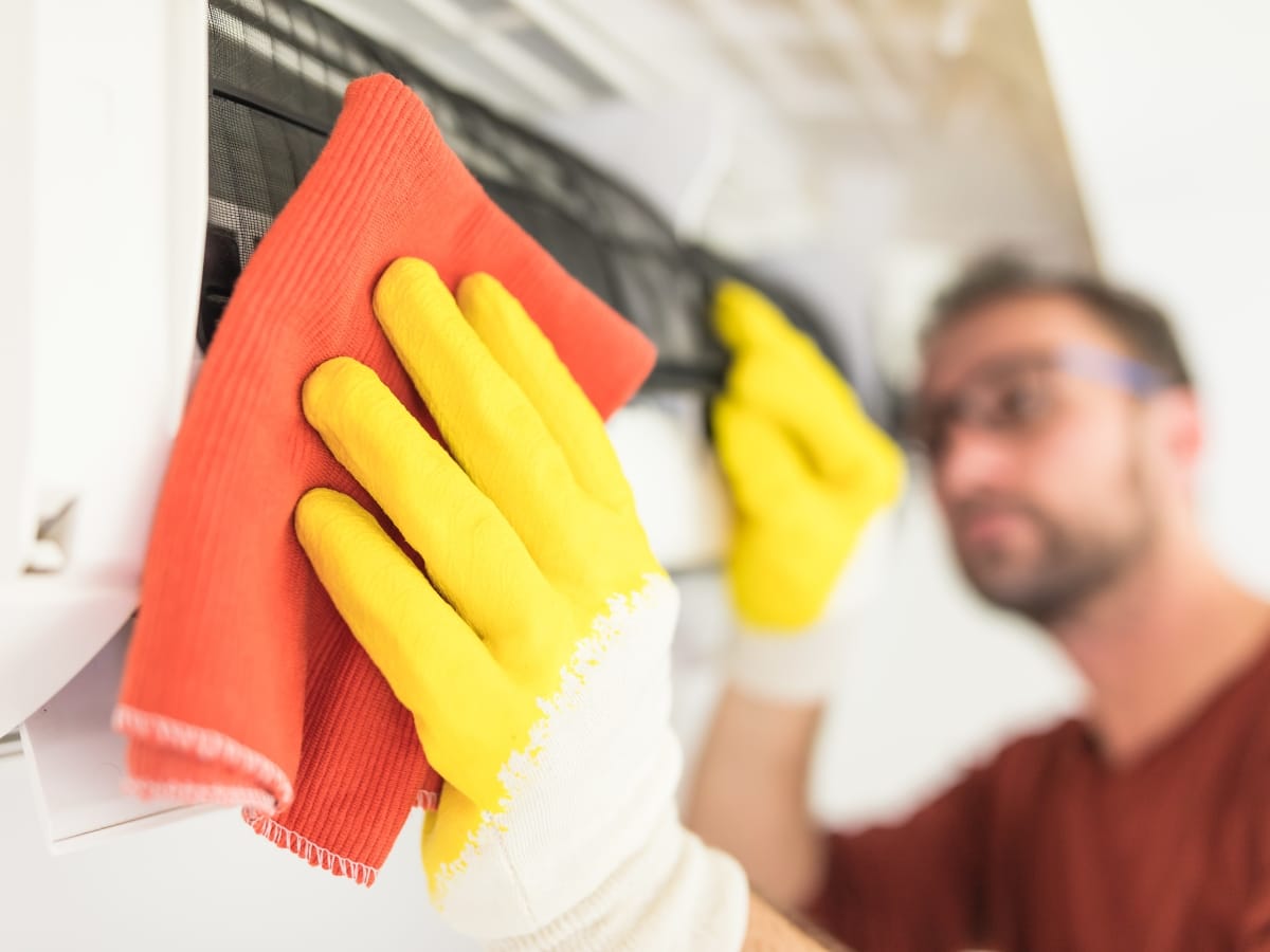 A person wearing yellow gloves performs AC filter cleaning, wiping the air conditioner filter with an orange cloth.