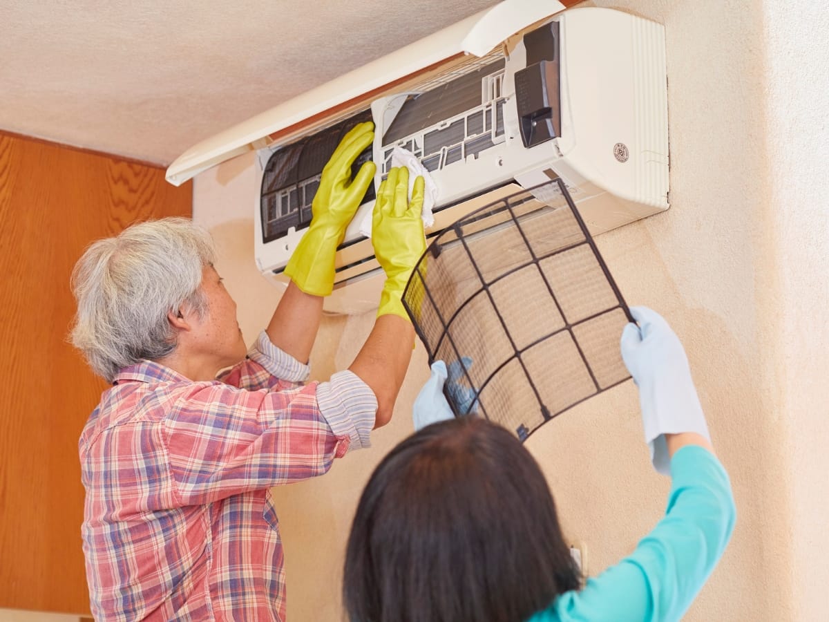 Two people wearing gloves perform AC filter cleaning as they clean the filters and interior of a wall-mounted air conditioning unit.