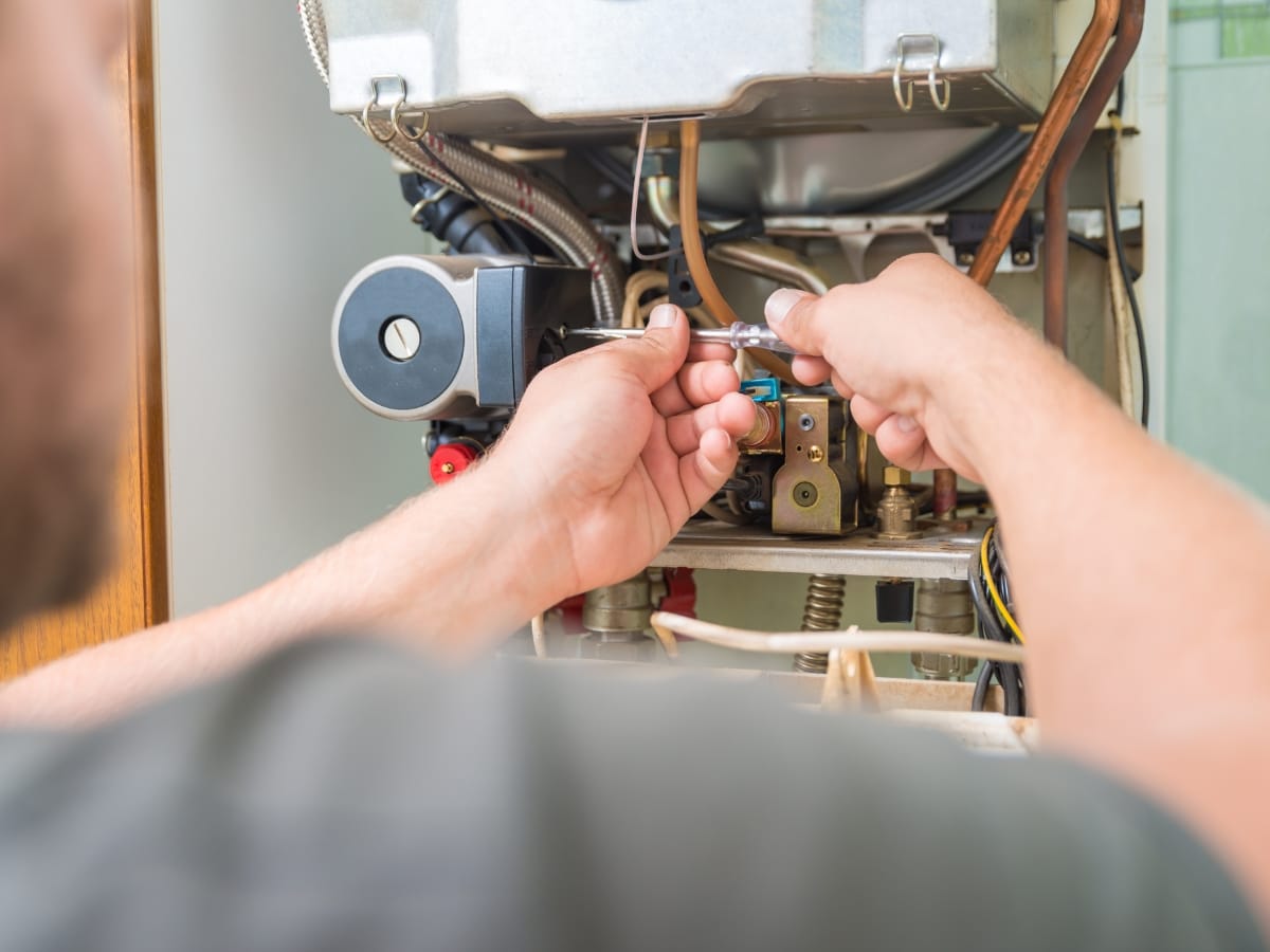A person uses a wrench to repair or adjust components inside an open gas boiler, focusing on areas near the pilot light or heating system.