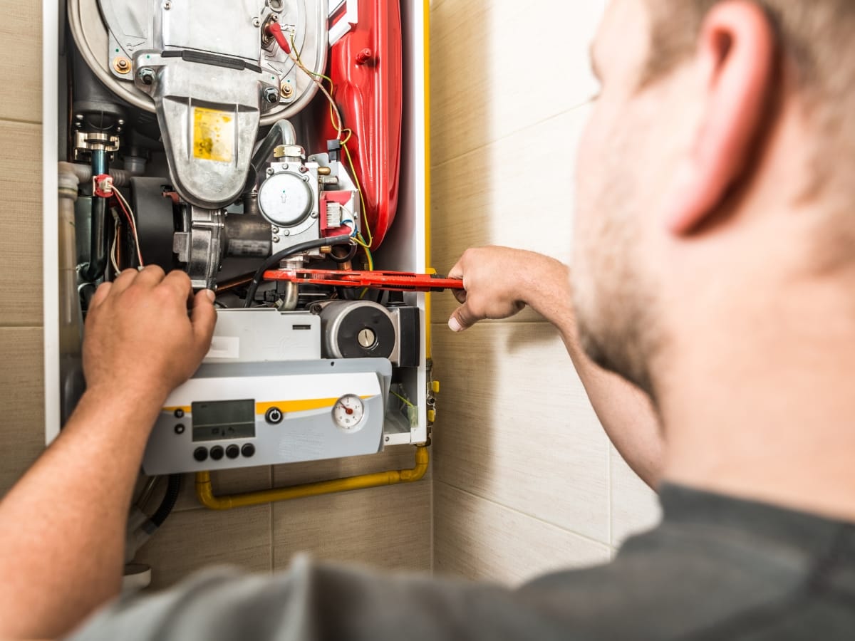 A person uses a wrench to adjust components inside an open gas boiler mounted on a wall, ensuring the pilot light is operating safely.