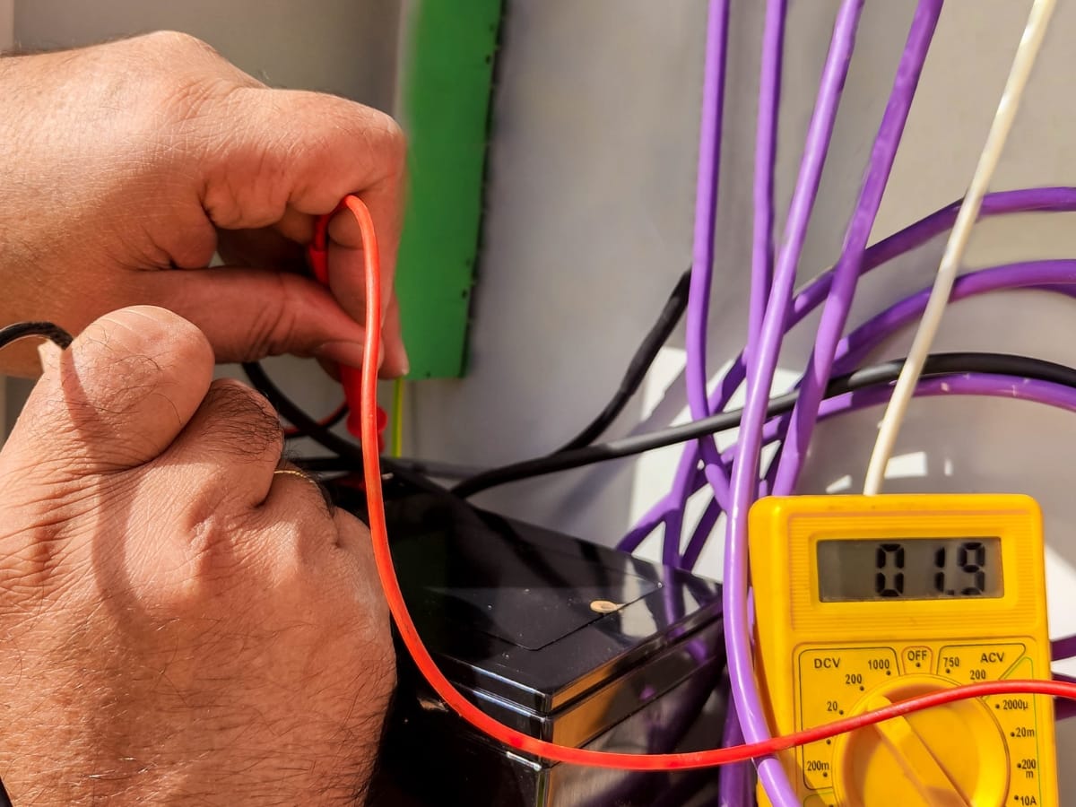 A person uses a digital multimeter to measure voltage on a black device with multiple colored wires visible, checking the pilot light circuit; the display shows 0.19 volts.