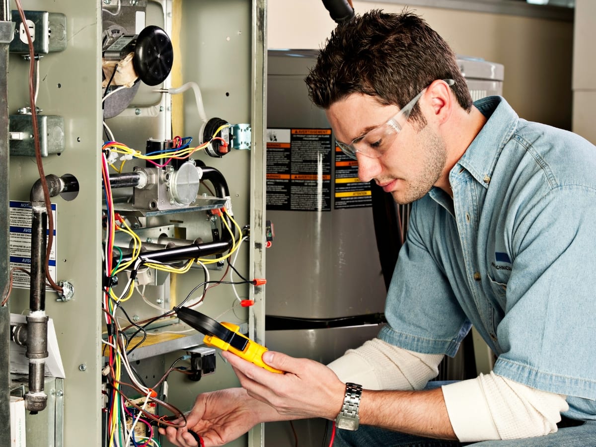 A technician wearing safety glasses uses a multimeter to test electrical wires and checks the pilot light inside an open HVAC unit.