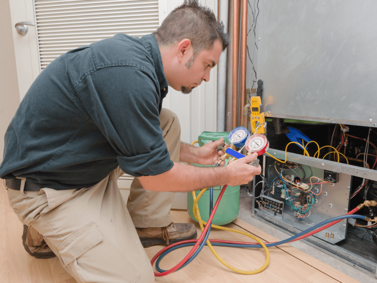 A technician uses gauges and colorful hoses to check and service an air conditioning HVAC unit, with wiring and tools visible around the open panel.
