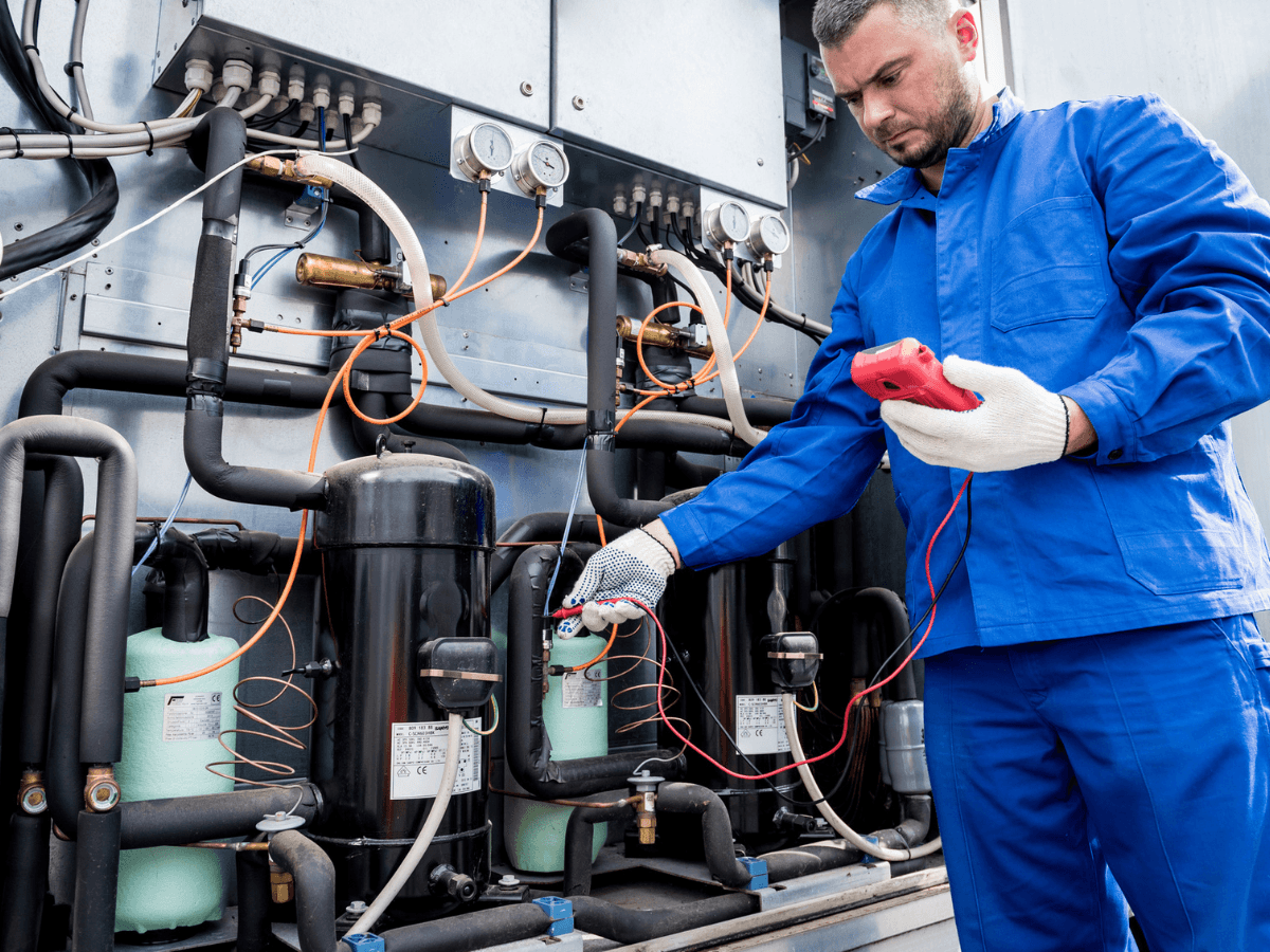 A technician in blue coveralls uses a handheld device to check the wiring of industrial refrigeration and air conditioning units.