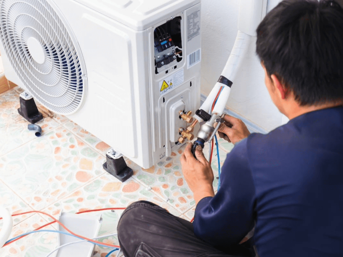 A technician in a navy shirt services an outdoor air conditioning unit, using tools to adjust wiring and connections.