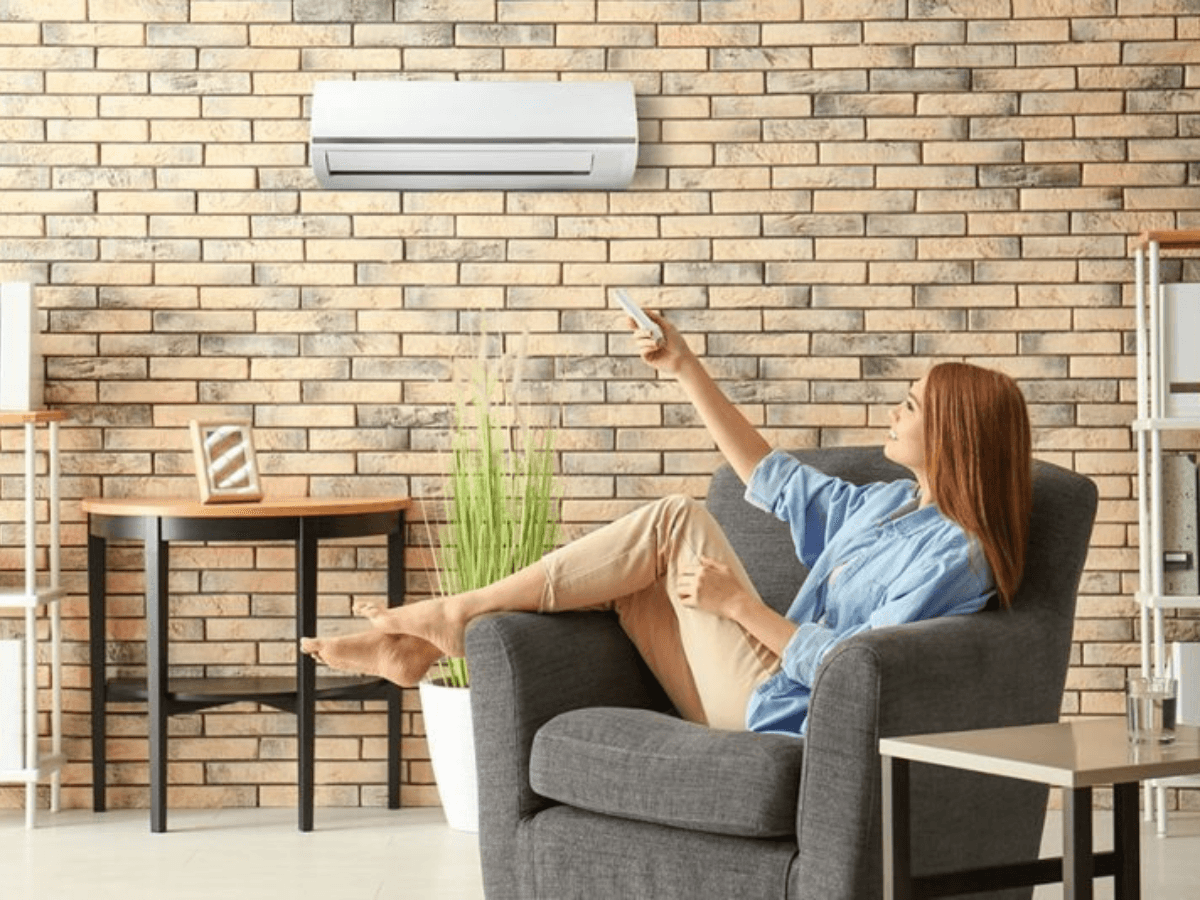 Woman sitting in an armchair points a remote control at a wall-mounted air conditioning unit in a living room with brick walls and modern decor.