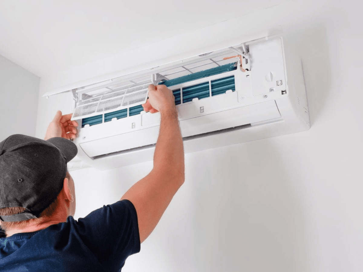 A person in a cap is inspecting or repairing the filter of a wall-mounted air conditioning unit.