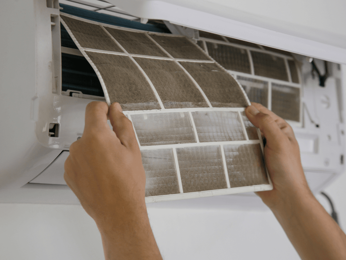 A person removes a dirty air filter, similar to furnace filters, from a wall-mounted air conditioning unit for cleaning or replacement.