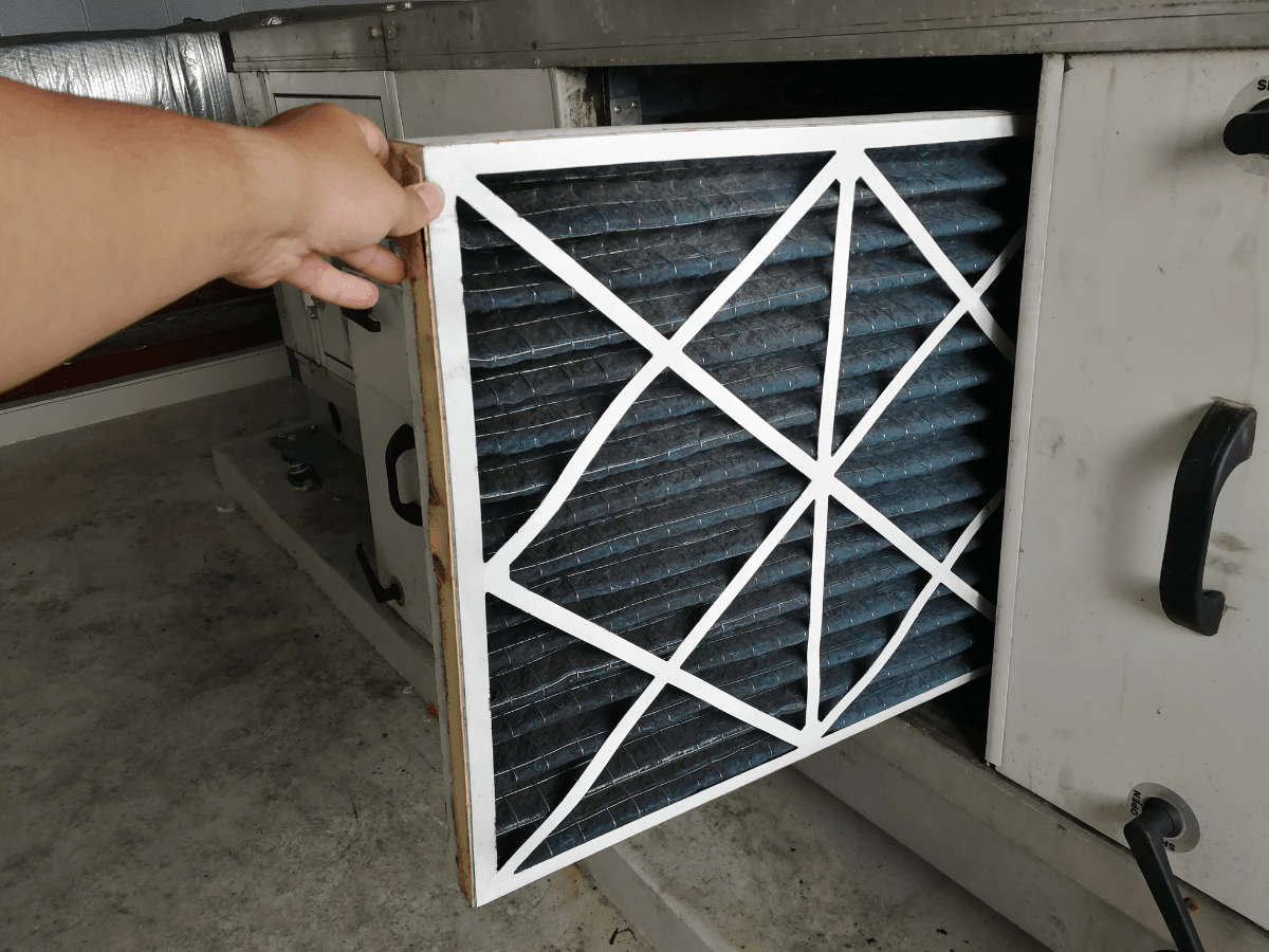 A hand removing a dirty furnace filter from a ventilation unit inside a building.