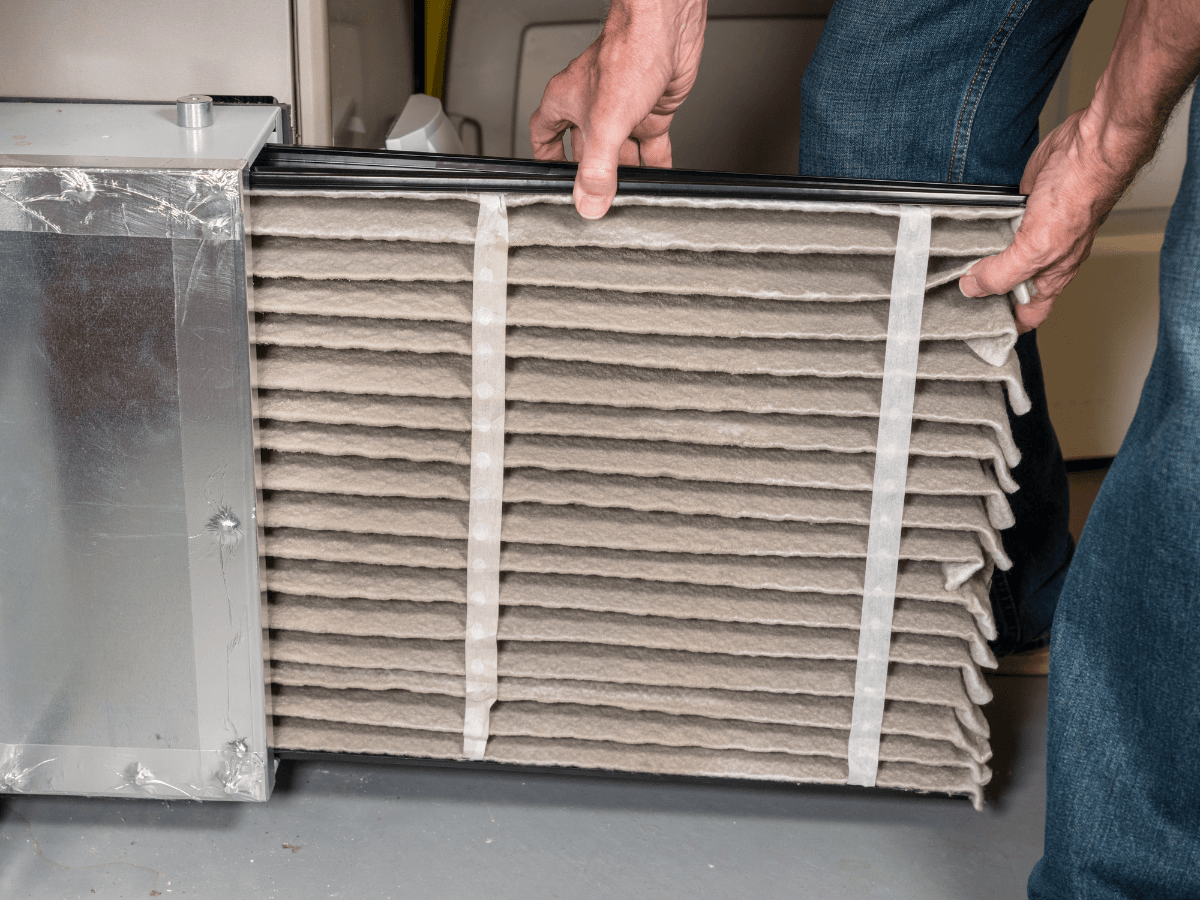 A person removes a dirty furnace filter from a residential HVAC system, revealing dust and debris on the filter surface.