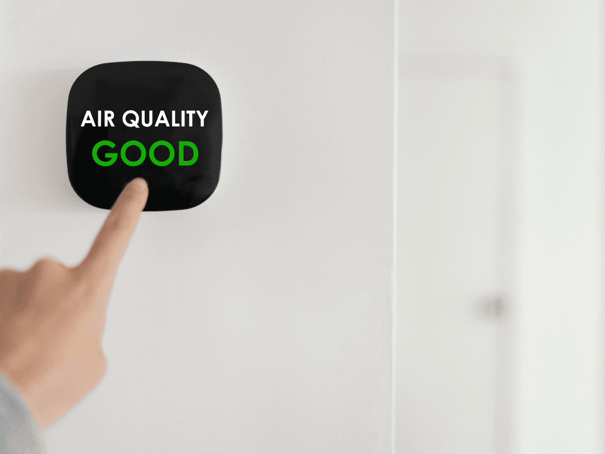 A person’s finger points to a digital air quality monitor on a white wall displaying the words "AIR QUALITY GOOD" in white and green text, highlighting the effectiveness of clean furnace filters.