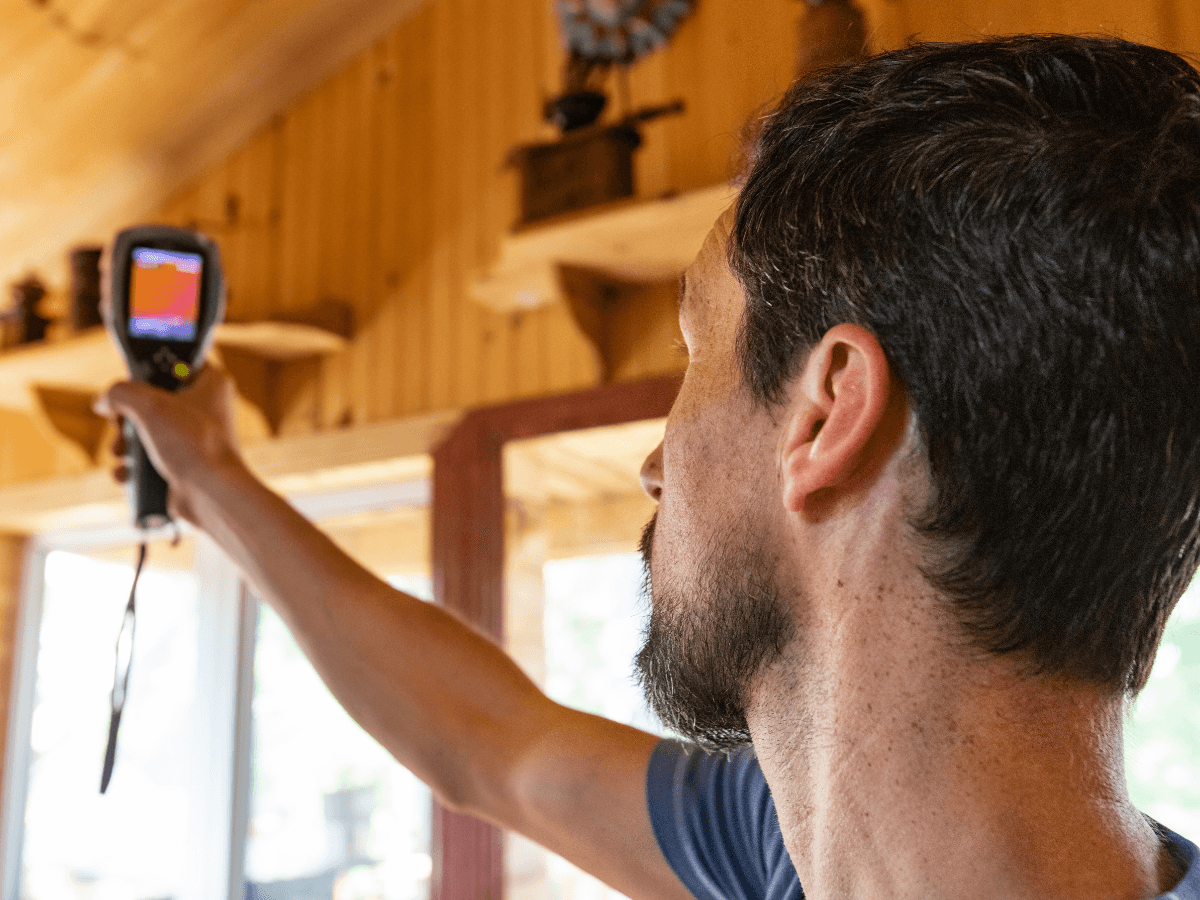 A person uses a handheld thermal imaging camera to scan the interior wall of a wooden room, checking for heat loss and inspecting areas near furnace filters.