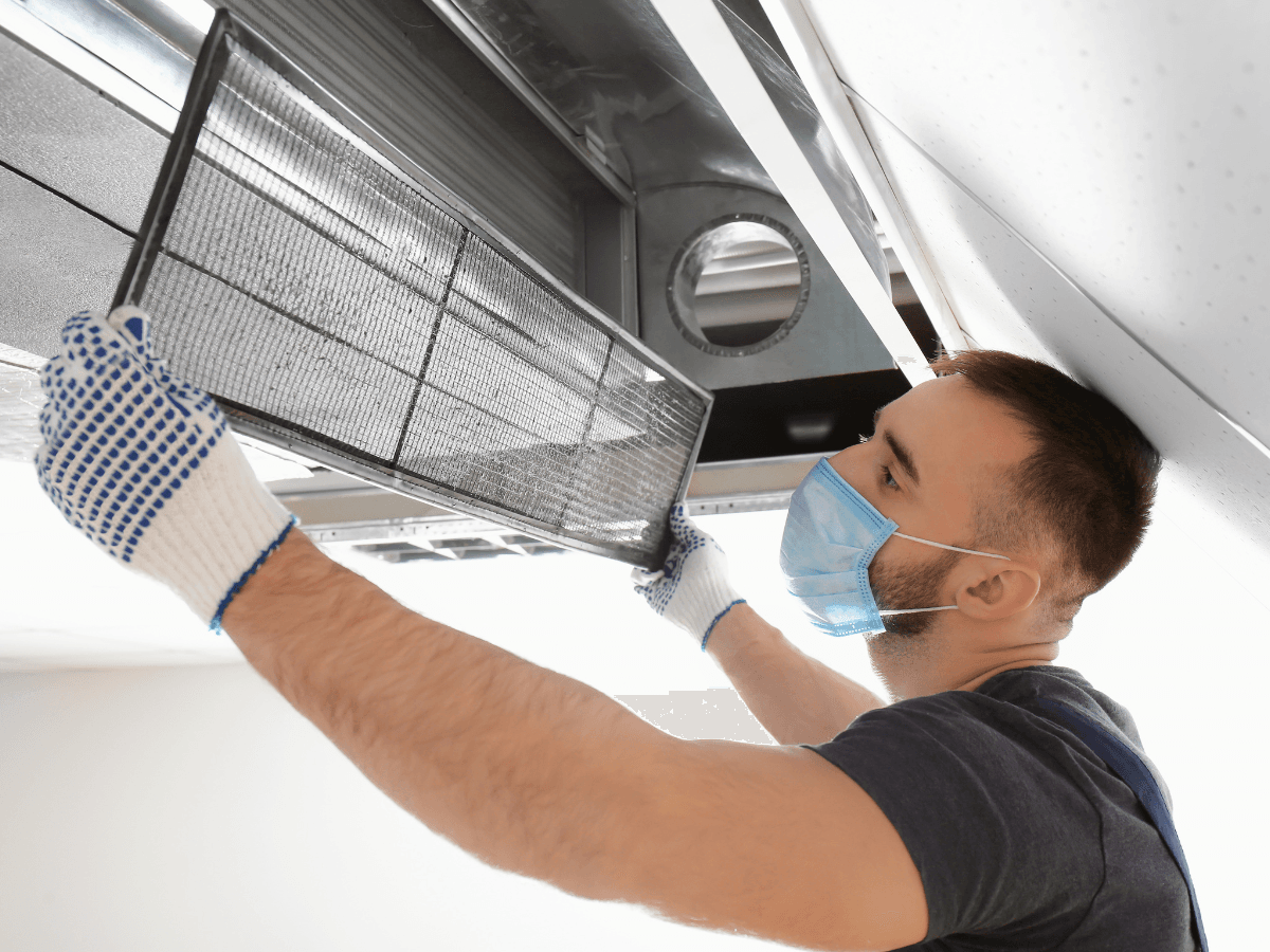 A man wearing gloves and a face mask removes an air filter, similar to furnace filters, from a ceiling ventilation duct for maintenance.