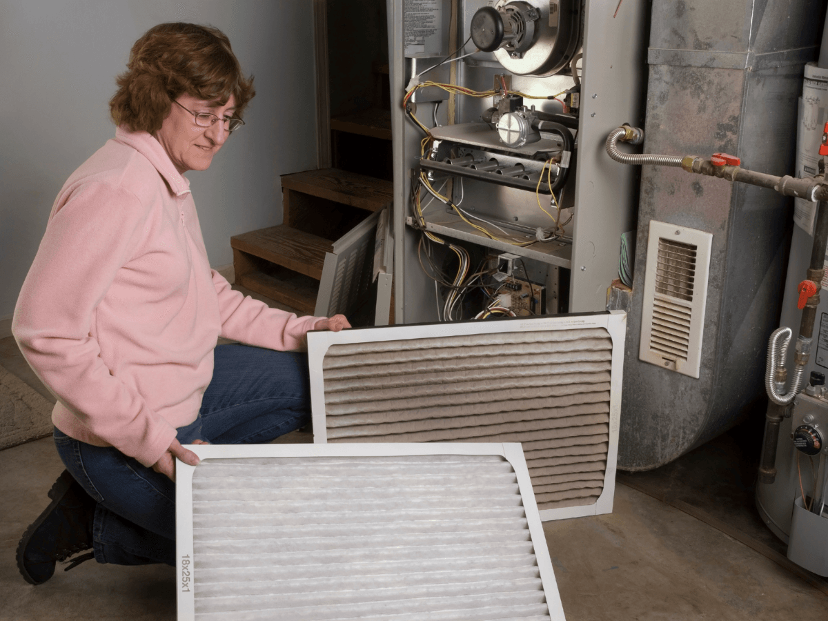 A woman compares clean and dirty furnace filters while kneeling next to an open furnace in a basement.