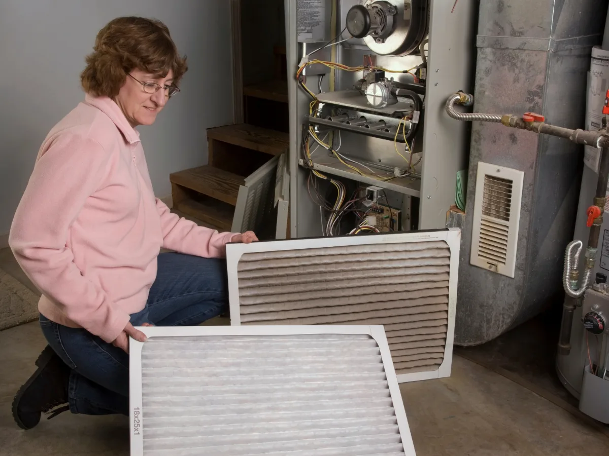 A woman compares clean and dirty furnace filters while kneeling next to an open furnace in a basement.
