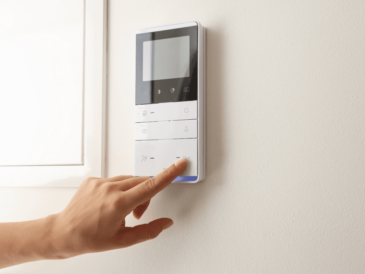 A hand presses a button on a wall-mounted digital home security or intercom control panel with a blank screen, perfect for summer AC prep and smart home convenience.