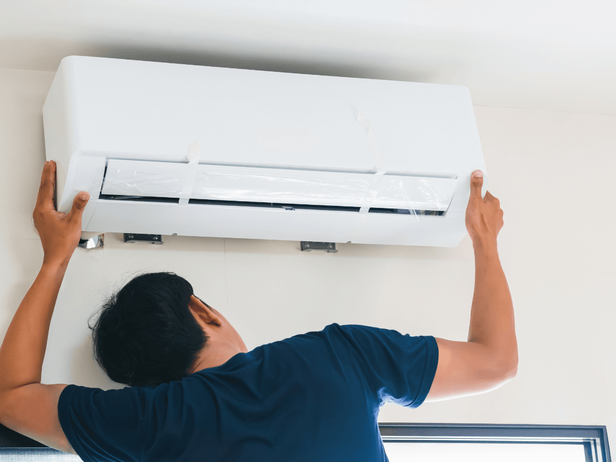 A person installs a wall-mounted air conditioning unit in a room, adjusting it to fit securely on the wall brackets as part of their summer AC prep.