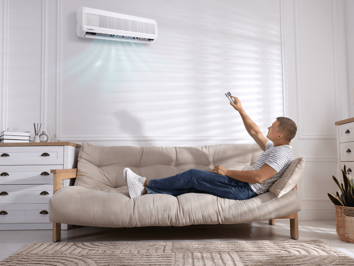 A man lies on a sofa using a remote to operate a wall-mounted air conditioner in a bright living room, enjoying the comfort of summer AC prep.