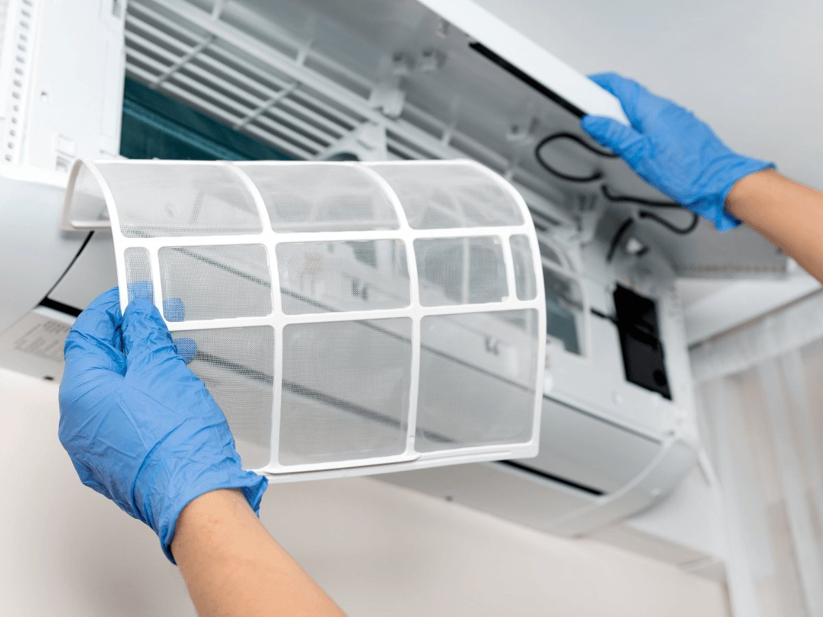 Person wearing blue gloves removes an air filter from a wall-mounted air conditioning unit as part of essential summer AC prep for cleaning or maintenance.