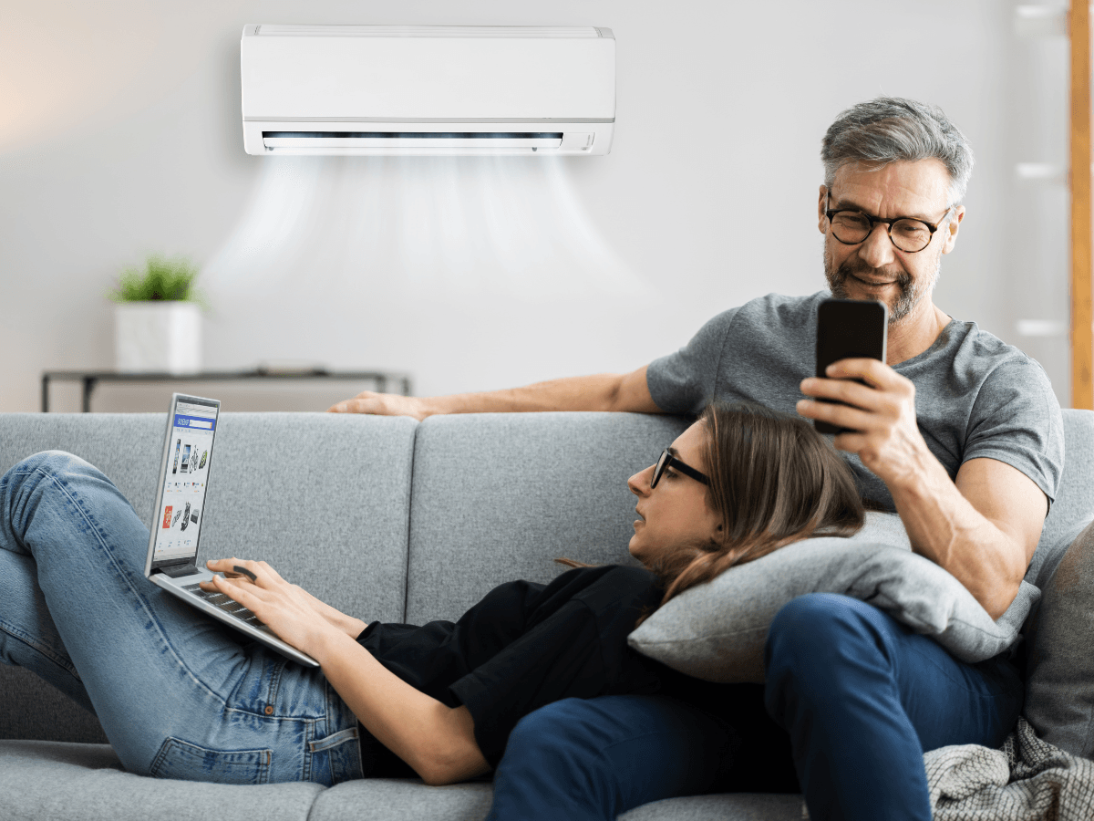A man and woman relax on a couch using a smartphone and laptop, enjoying cool air from a wall-mounted unit—a perfect scene of summer AC prep.