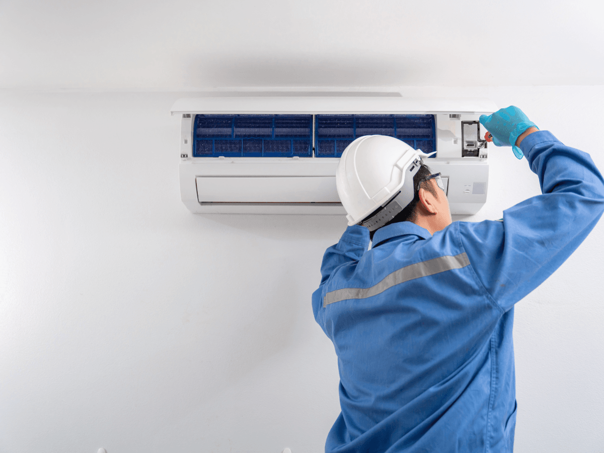 A technician in a hard hat and blue uniform performs summer AC prep as he repairs or maintains a wall-mounted air conditioning unit.