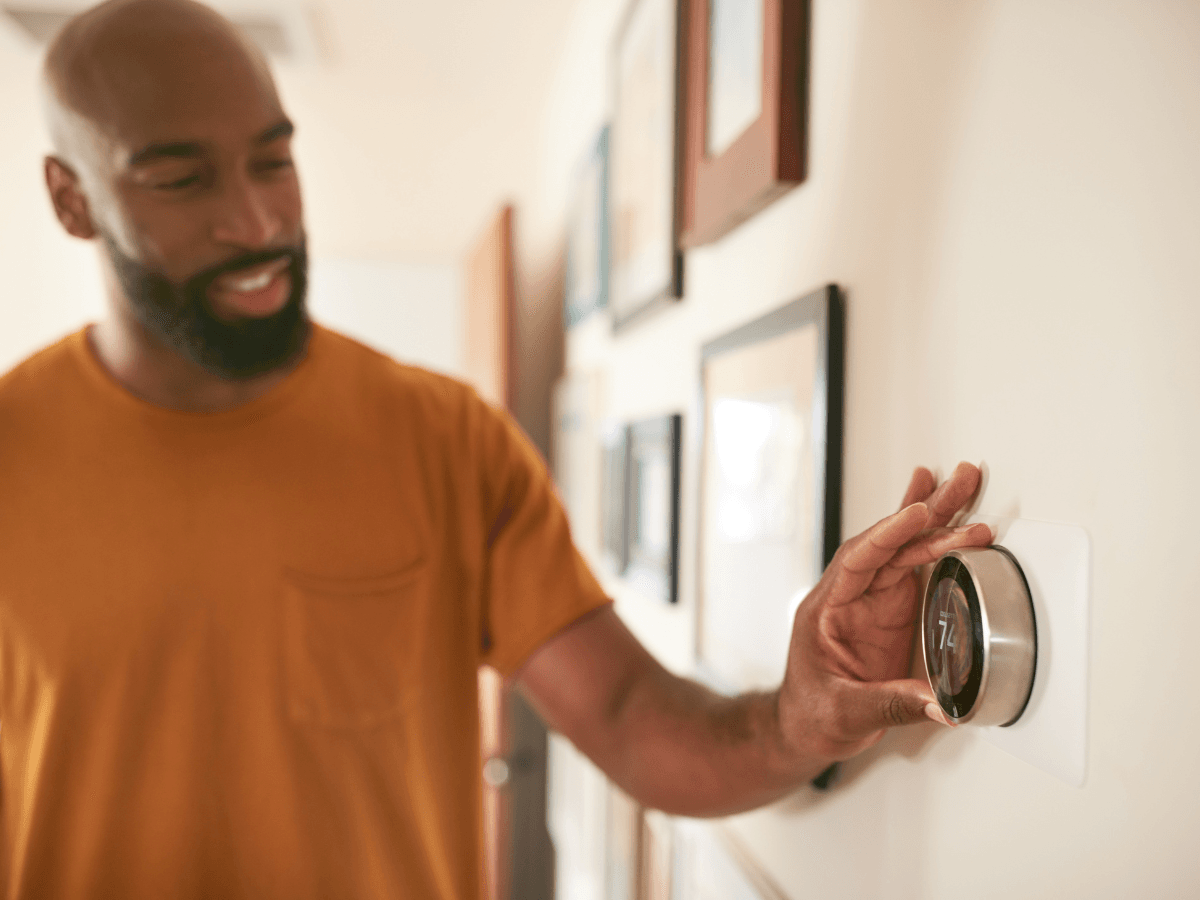 A man in an orange shirt adjusts a round smart thermostat on a wall decorated with framed pictures, trying to figure out why the thermostat is not working.