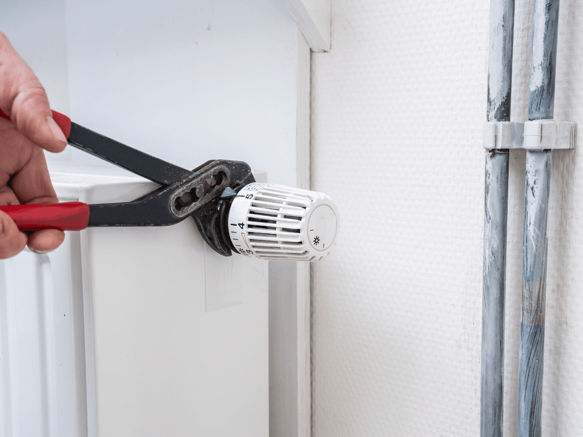 A person uses a wrench to tighten or adjust a valve on a white radiator next to exposed pipes in a room with white walls, possibly addressing an issue with the thermostat not working.