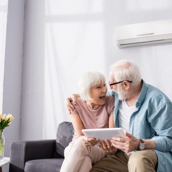 An older couple sitting on a couch looking at a tablet computer enjoying .