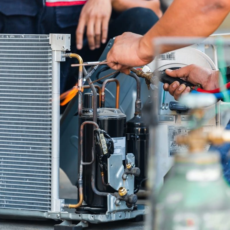 Two technicians diligently work on an air conditioning unit, using tools to connect wires and components. Their expertise extends beyond cooling systems; they're trained to spot signs your furnace needs repair, ensuring every aspect of your comfort is addressed with precision.